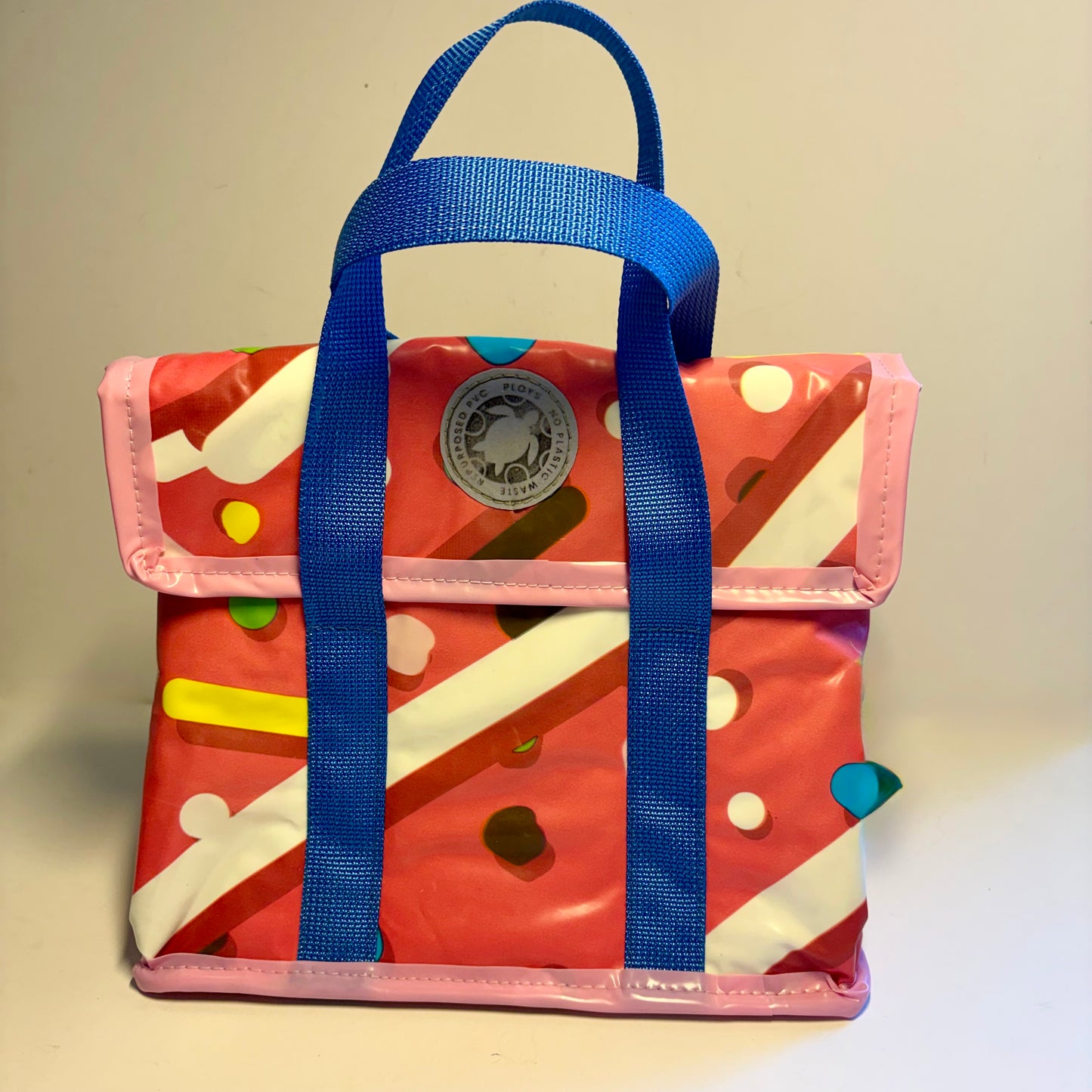 A small glossy pink lunch bag with blue handles, featuring a donut themed print with white stripes, colourful dots, and velcro closing front flap.