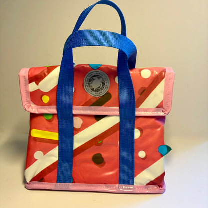 A small glossy pink lunch bag with blue handles, featuring a donut themed print with white stripes, colourful dots, and velcro closing front flap.