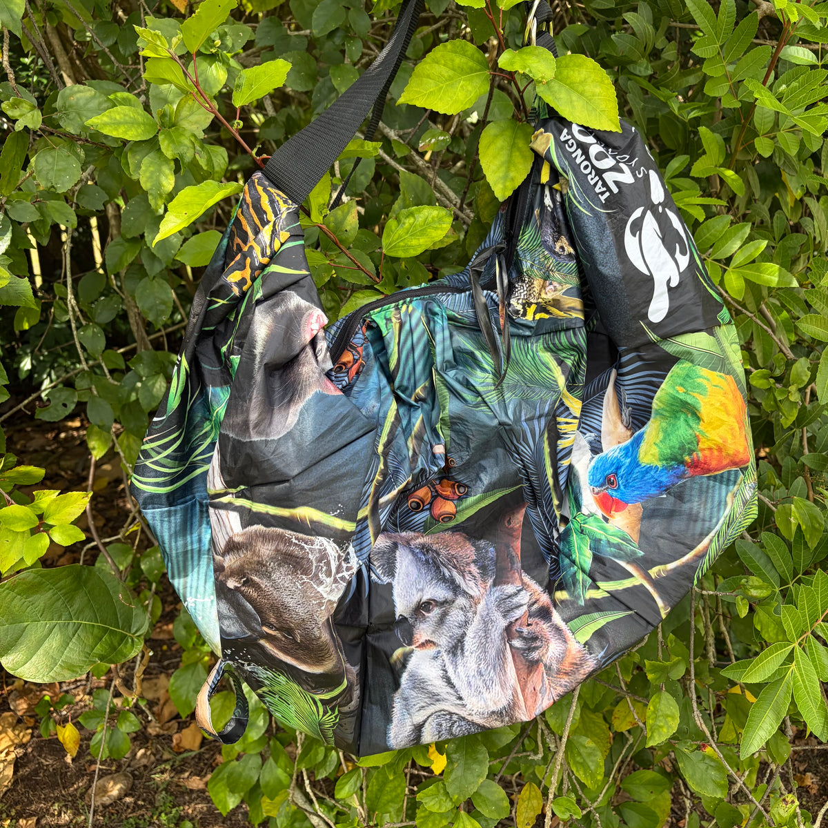 A black zippered Tasuki duffel bag made from upcycled umbrellas with a colourful Australian nature animal and jungle print, hanging outdoors among green leaves. The bag features a black shoulder strap and a white logo on the side.