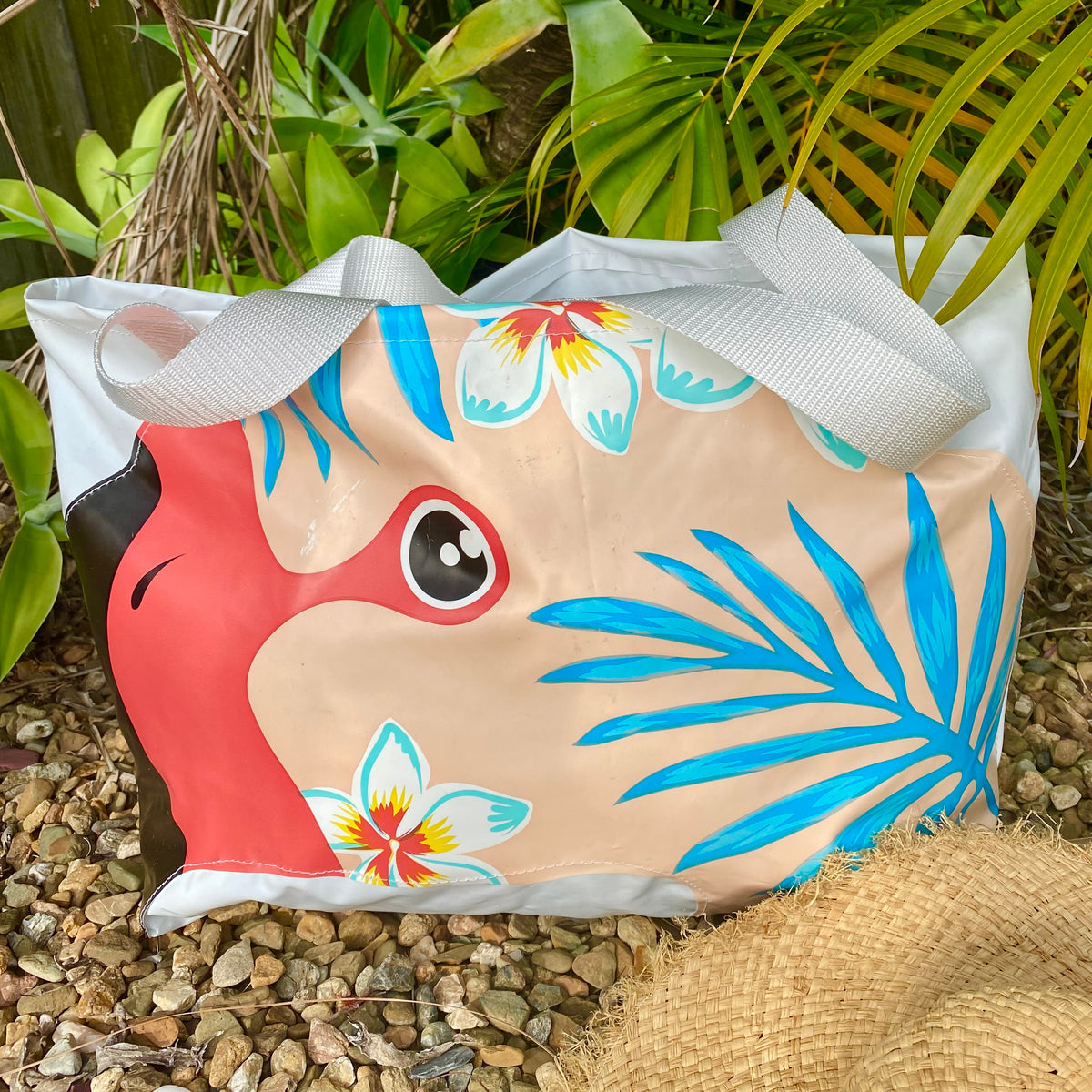 A beach tote bag made from upcycled pool inflatables with a tropical design featuring a red flamingo face, blue palm leaves, and white flowers on a beige background, with white handles.